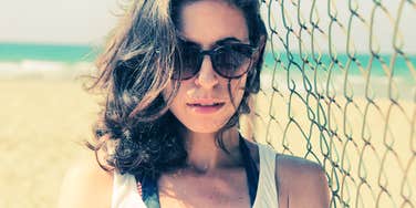 Woman wearing sunglasses standing near a chain-link fence at the beach.