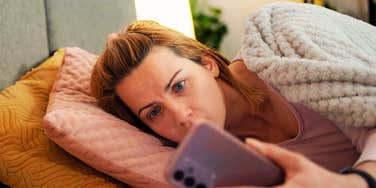 tired woman lying in bed under a blanket at night staring at her phone screen with wide, anxious eyes.