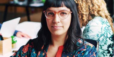 confident professional woman with glasses making direct eye contact in modern workspace, poised expression and calm presence conveying strong communication skills and natural authorit