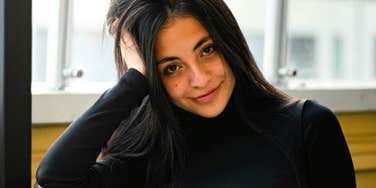 A close-up portrait of a woman with long dark hair sitting indoors near a window.