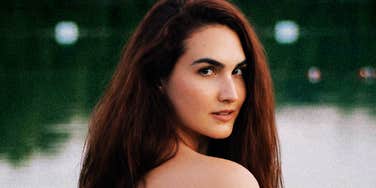 A close-up portrait of a woman with long brown hair looking over her shoulder outdoors.