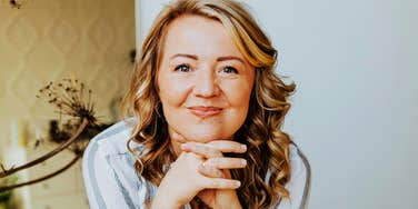 smiling middle-aged woman with shoulder-length wavy blonde hair resting her chin on her hands, soft natural light, calm indoor setting