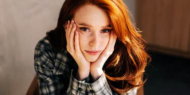 thoughtful red-haired woman resting her face in her hands, intense introspective gaze in warm natural light.