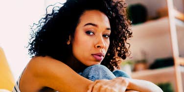 close-up of pensive young woman with curly dark hair sitting on the floor hugging her knees, soft natural indoor light