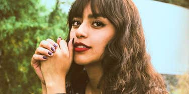 A close-up portrait of a woman with long wavy hair looking toward the camera outdoors.