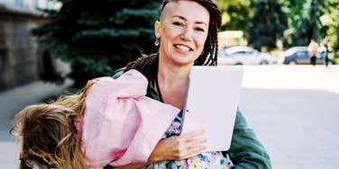 smiling working mom holding a laptop while carrying her tired child outdoors, multitasking between career and motherhood
