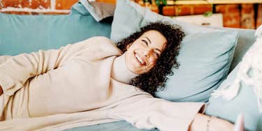 woman relaxing alone on a cozy couch, smiling peacefully in a sunlit living room, calm and content energy 