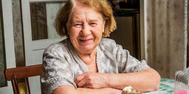A kind grandmother smiling warmly at a dining table, illustrating the moment of sharing life-changing wisdom and family legacy through a simple conversation.