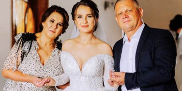 Happy parents hold the hands of the bride who is getting married and support her, wearing a wedding dress and a long veil.