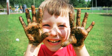 Little boy with muddy hands smiling and holding them up, illustrating the high-energy chaos parents of active young boys know well