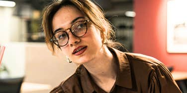 A capable professional woman in a brown shirt sitting at her office desk with a curious expression, representing the 'missing personality trait' that often holds back high-performers.