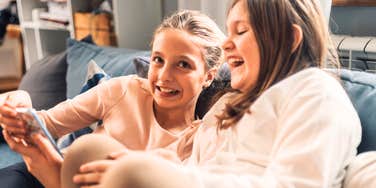 Two sisters are sitting on a sofa, enjoying funny content on a smartphone, creating a joyful and connected atmosphere