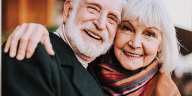 A joyful husband and wife in their sixties sharing a warm embrace, illustrating the authentic happiness that comes from decades of navigating the realistic and unglamorous aspects of partnership.