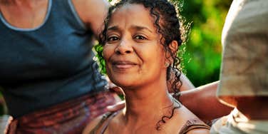 A mature woman with a warm, radiant smile sitting outdoors at a wellness retreat, embodying the open and welcoming body language that instantly puts others at ease.