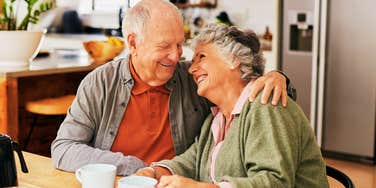 A happy senior couple sharing a tender embrace and laughing over coffee, illustrating the deep affinity and personal transformation that comes from decades of a successful marriage.