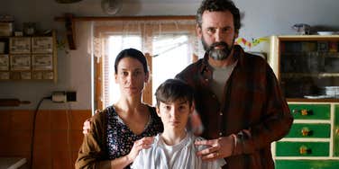 A tired middle-class family standing together in a kitchen with their child, illustrating the 'dual-income trap' and the systemic pressures on the modern American family.