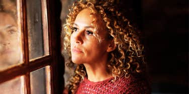 A thoughtful adult woman looking out a window with her reflection visible in the glass, illustrating the 'internalized habits' and subtle daily choices that lead to chronic unhappiness.