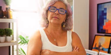 A confident Hispanic woman in her 50s or 60s with gray hair and glasses, standing with arms crossed to represent the self-assurance and lack of apologies that come with middle age.
