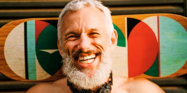 A mature man with grey hair laughing while holding a skateboard outdoors, illustrating the 'functional fitness' and playful habits that keep people feeling decades younger after fifty.