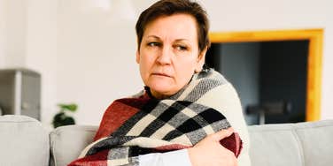 Middle-aged 50s woman seated on sofa in living room covered with warm plaid blanket