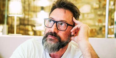 In a quiet room at home, a 50-year-old man with glasses sits alone, his face marked by sadness and concern, as he struggles with feelings of loneliness, emotional exhaustion, and personal worries