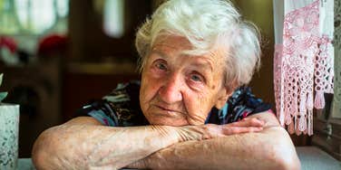 A portrait of a gray-haired elderly woman sitting near a window, illustrating the 'cognitive dissociation' and dual-reality perception common in dementia.
