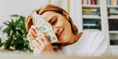 A woman with a joyful expression holding cash close to her face; illustrating the "strange but effective" habits people use to shift their vibration and attract more money into their everyday lives.