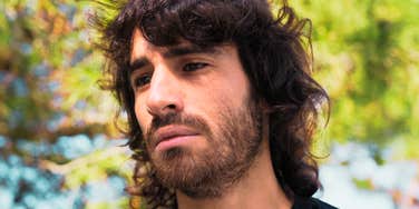 A somber man with long hair sitting alone in nature, appearing depressed and overwhelmed; illustrating the 'unseen' emotional struggle of men facing social disconnection in a modern landscape.