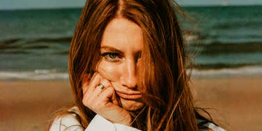 A woman with a sad expression stands on a scenic beach, illustrating the internal disconnect of feeling lonely even when life appears full on the surface.