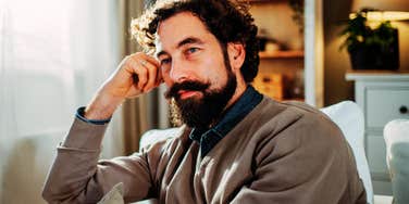 A thoughtful man sitting in a living room, looking away with a composed expression; a visual of the emotional self-regulation and focus characteristic of exceptionally resilient people.