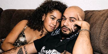 A couple sitting on a couch with the man looking thoughtfully at the camera, finger on his temple, while the woman looks at him; illustrating the complex reality of conflict that social media often masks.