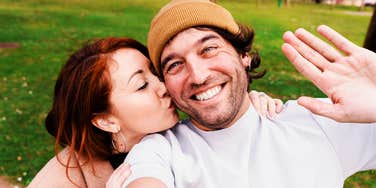 A joyful couple enjoying a day in nature, with the woman kissing the man’s cheek during a selfie; illustrating the spontaneous and consistent ways partners express their devotion and love every day.