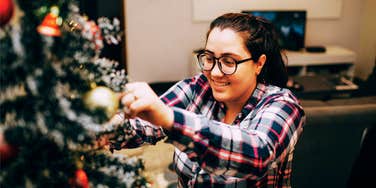 A woman in a casual shirt happily tending to a Christmas tree; illustrating the psychological connection between extended holiday celebrations and personal happiness.