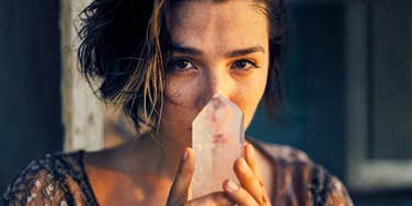 A woman holding a healing crystal close to her face with a guilty expression, illustrating the conflict between progress in therapy and the impulse to buy anxiety-relief crystals.