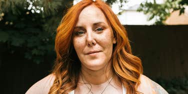 A portrait of a red-haired woman in her late 30s standing outdoors; a representation of the 'alarmingly common' state of high-functioning exhaustion that therapists identify as an inhumane way of living.