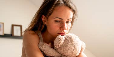 Lonely woman embracing plush teddy bear while sitting on bed indoors