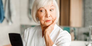 A portrait of a mature woman in a bathrobe preparing for a makeover in front of a mirror; illustrating the viral 'Glam-Ma' trend where eighty-year-olds reclaim their style and confidence through high-glamour transformations.