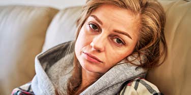 A woman sitting in solitude on a living room couch, looking depleted and overwhelmed; illustrating 'maternal burnout' and the systemic pressure on women to perform domestic happiness while struggling with internal depression.