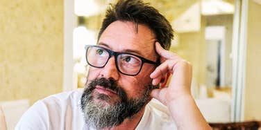 A 50-year-old man with glasses sitting alone in a quiet room, looking concerned and emotionally exhausted; representing the 'mid-life financial crisis' and the daunting reality of starting a retirement plan later in life.