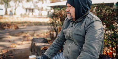 unhoused man sitting on sidewalk holding cup