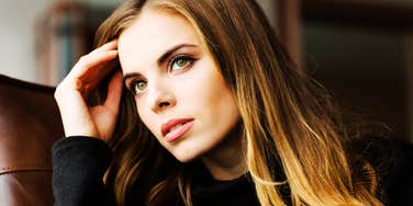 A young woman sitting on a sofa in a thoughtful, guarded pose; illustrating the 'selective vulnerability' and calculating stillness that often precede more obvious betrayals in personal relationships.