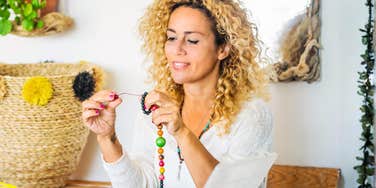 A woman sitting at a table making intricate jewelry with colorful beads; illustrating the therapeutic benefits of fine motor activities and how making tiny crafts can improve mental focus and physical wellbeing.