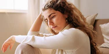 A woman sitting in a living room looking away with a sense of isolation; illustrating the 'silent erosion' of a relationship where communication has shifted from active engagement to habitual defensiveness and emotional distance.