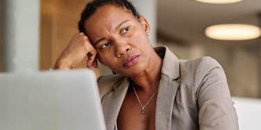 A businesswoman looking exhausted and defeated while sitting with her laptop; illustrating the deep fatigue and feeling of being 'unsafe' that comes with prolonged workplace stress and a shattered worldview.