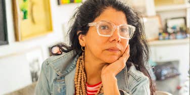 A Latin woman in her 40s or 50s attempting to work from home while appearing visibly drained; illustrating the 'double burden' of unpaid labor and professional demands that characterizes the Gen-X female experience in 2026.