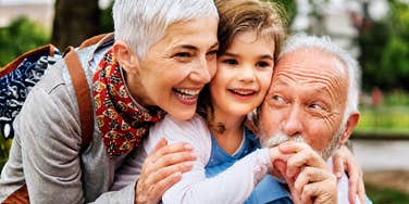 A close-up portrait of smiling grandparents embracing their granddaughter outside, capturing the stabilizing influence of grandparent attachment after early childhood disruption.