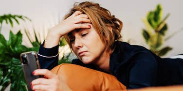 A candid side view of an exhausted woman lounging on a sofa in loungewear, completely absorbed in her smartphone screen; illustrating the 'bed rotting trend' and the generational rejection of hyper-productivity.