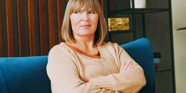 A close-up portrait of a displeased mother in her 50s sitting in a living room with a folded, defensive posture; illustrating 'the leisure guilt disconnect' where generations clash over productivity and rest while traveling.