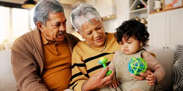 A close-up lifestyle shot of a smiling grandmother and grandfather bonding with a toddler over toys inside a house; illustrating 'the care economy imbalance' where historical family support structures collide with modern professional life.