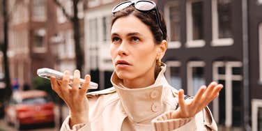 A young woman in a beige trench coat looking up at the sky in complete disbelief while holding her smartphone horizontally; a visual representation of 'the boundaried response' and Gen-Z communication exhaustion.